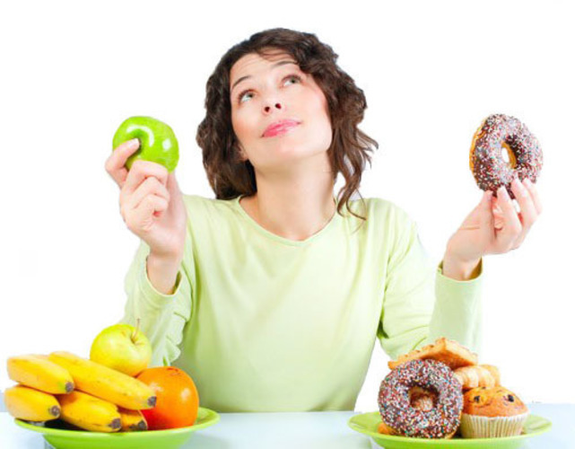 Žena sedící u stolu s ovocem a pečivem a držící jablko a koblihu 